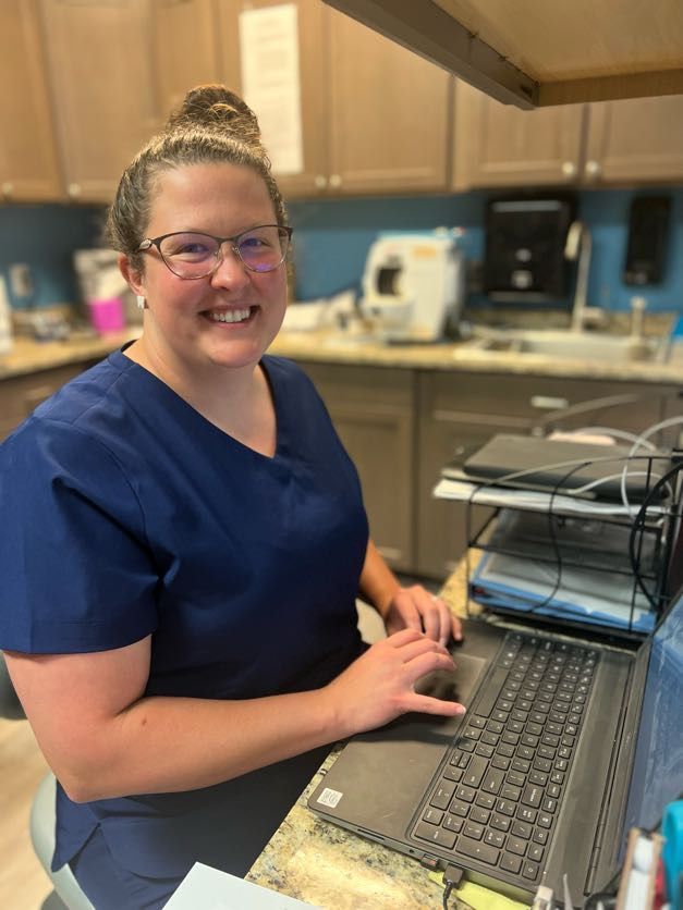 Woman in blue scrubs smiling, typing on a laptop in an office setting.