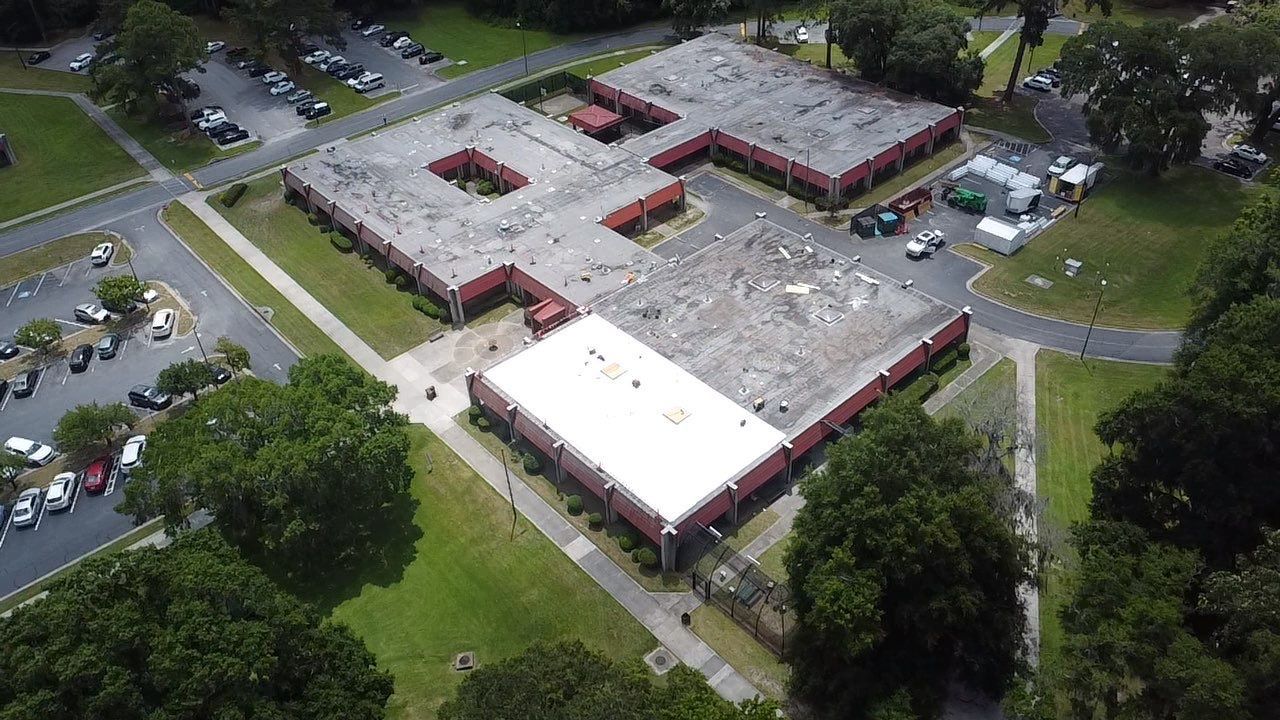 Aerial view of a red brick building with a white roof, surrounded by trees and parking lots.