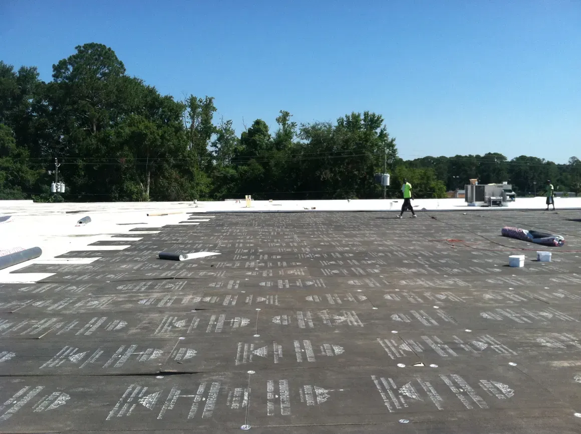 Workers on a flat roof, laying down white roofing material. Trees in the background, blue sky.