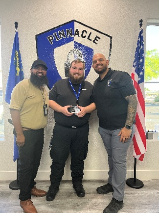 Three men pose in front of a Pinnacle logo. The central man holds an object. Flags are on either side.