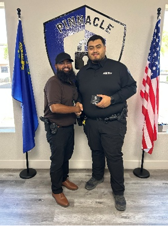 Two men shaking hands, one in brown shirt, the other in security uniform, in front of a logo and flags.