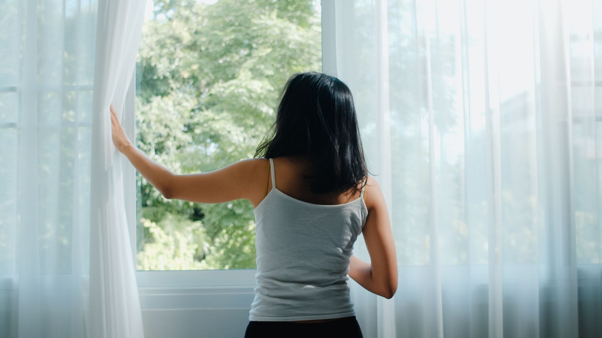 Woman in a tank top opening sheer curtains to look out a window with a green tree view.