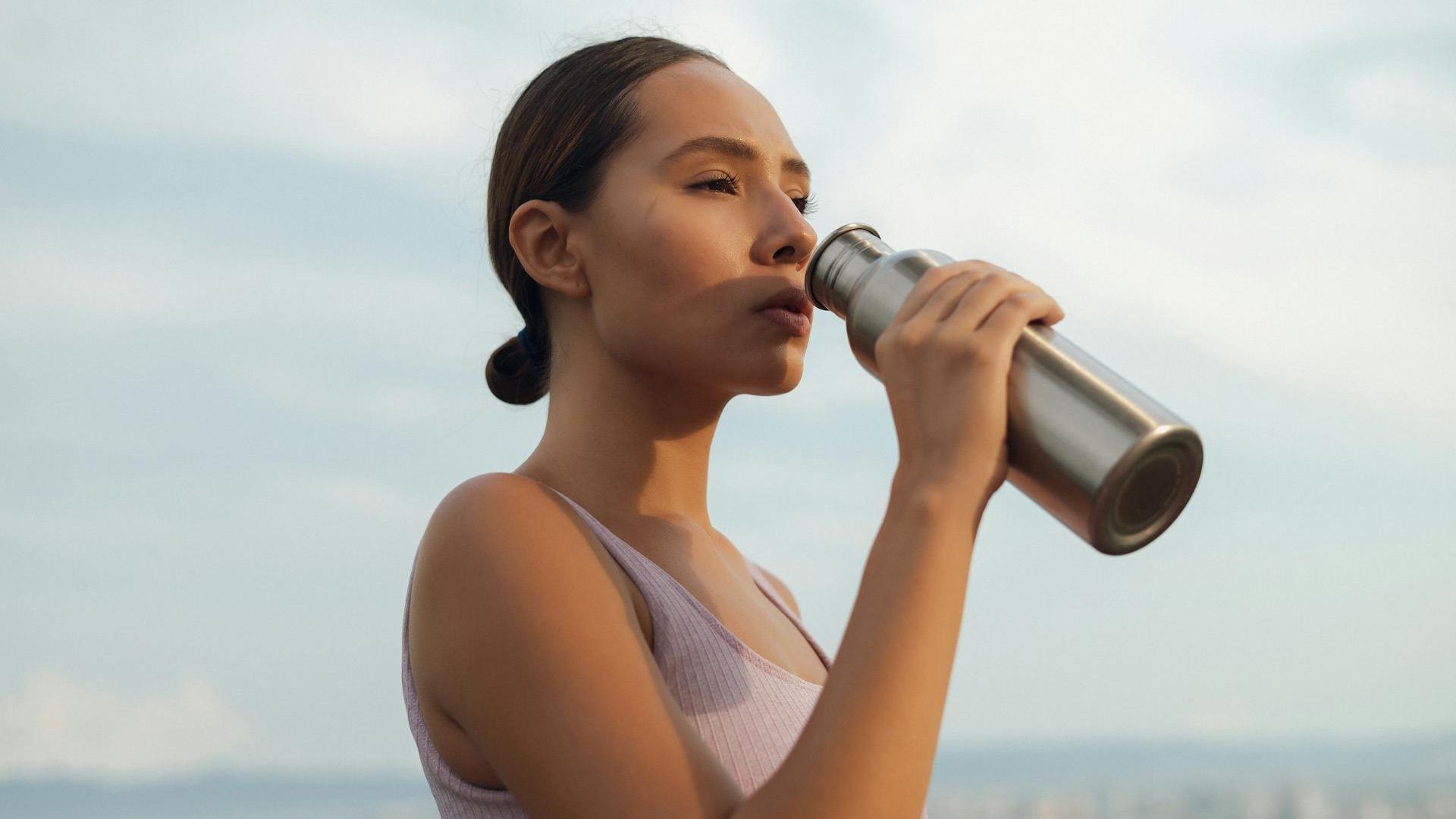 Woman in activewear drinks from a metal water bottle outdoors, lit by sunlight, with a bright sky in the background.
