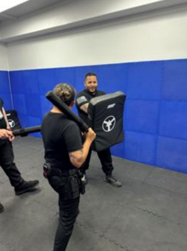 Two people practicing self-defense techniques, one with a baton, the other holding a shield in a training room.