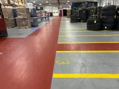 Warehouse aisle with red walkway, yellow floor markings, and stacked pallets and coiled materials on the right.