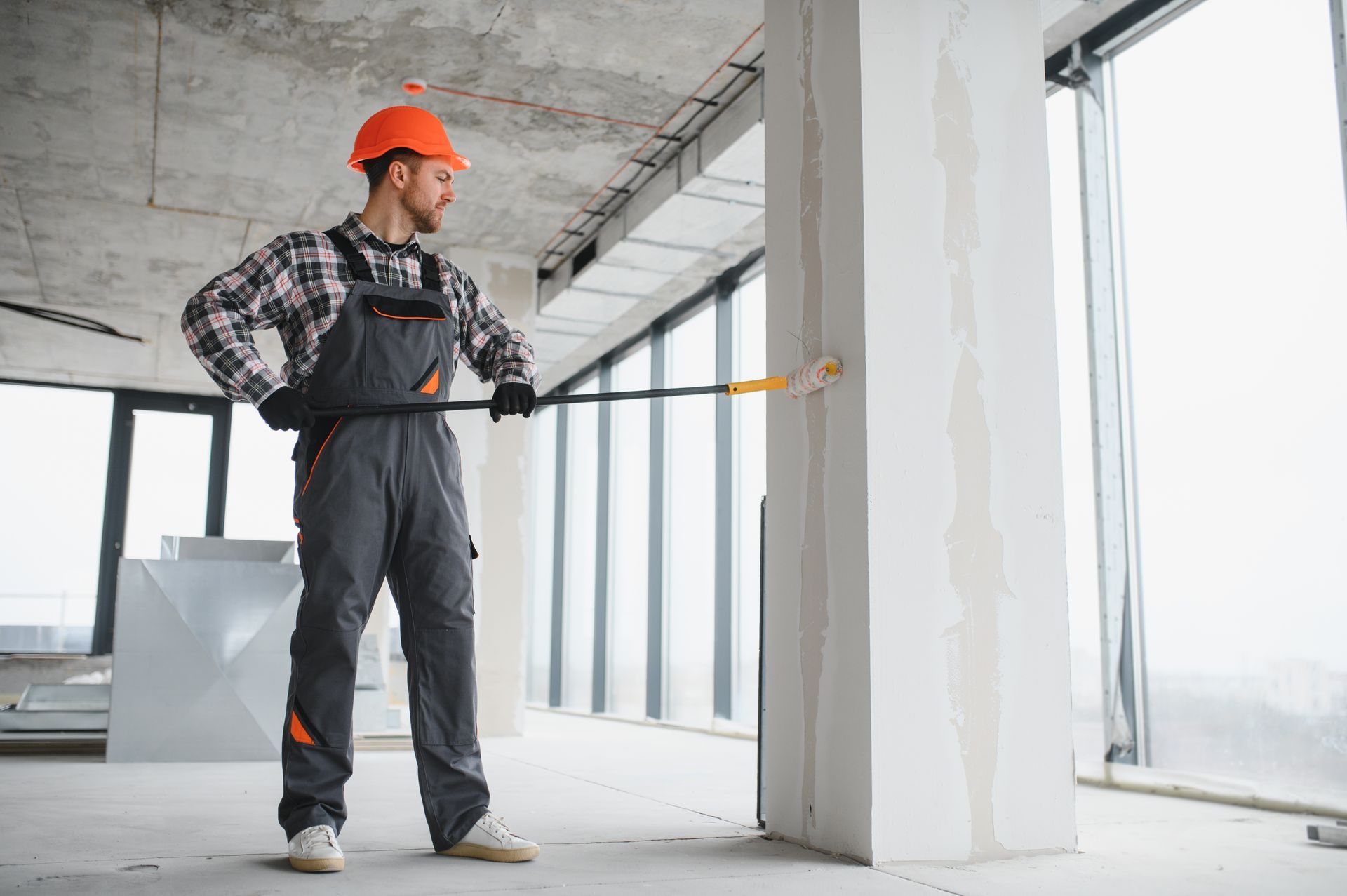 Construction worker using a paint roller to coat a white wall in an unfinished room