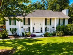 White suburban house with black shutters, white columns, and a bright green lawn under blue sky