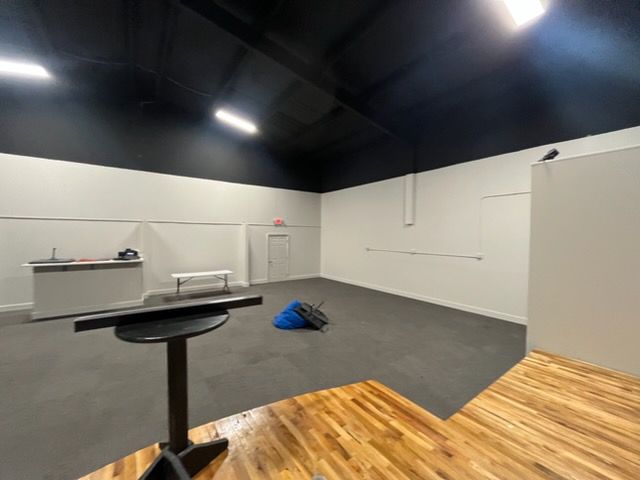 Empty indoor room with dark ceiling lights, gray floor, white walls, and a wooden platform in the foreground