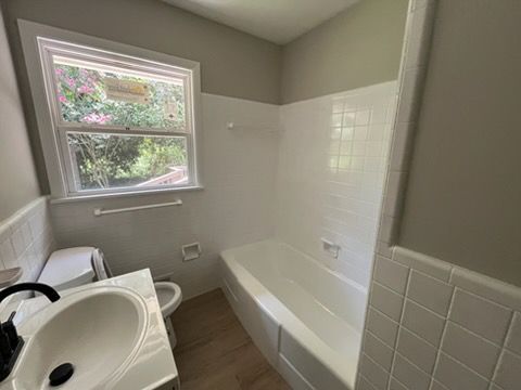 Small bathroom with white tub, sink, toilet, and window with greenery outside.