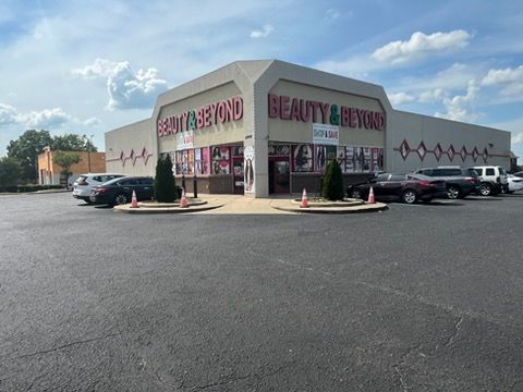 Beauty & Beyond storefront with pink sign, parked cars, and a wide asphalt parking lot under a blue sky.