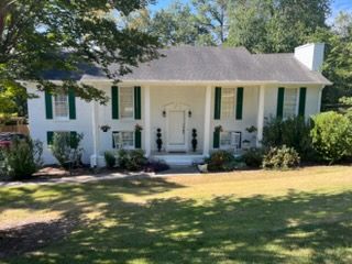 White house with green shutters, front porch, and large front lawn under trees.