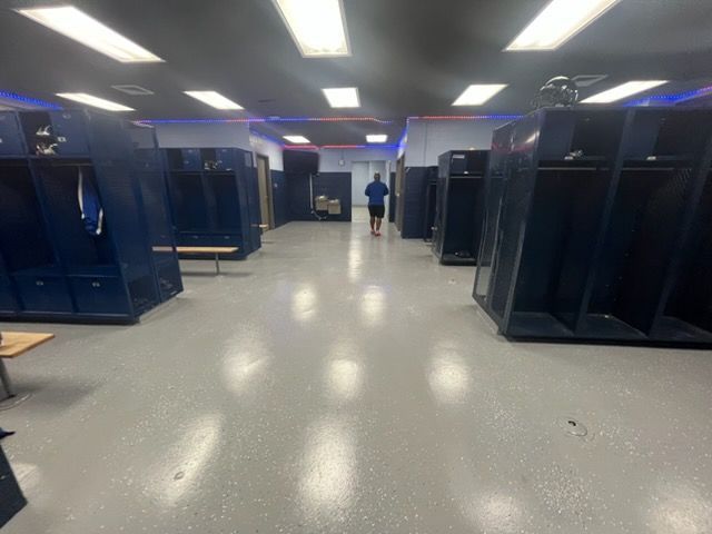 Spacious locker room with blue lockers and bright ceiling lights; a person stands in the distant center.