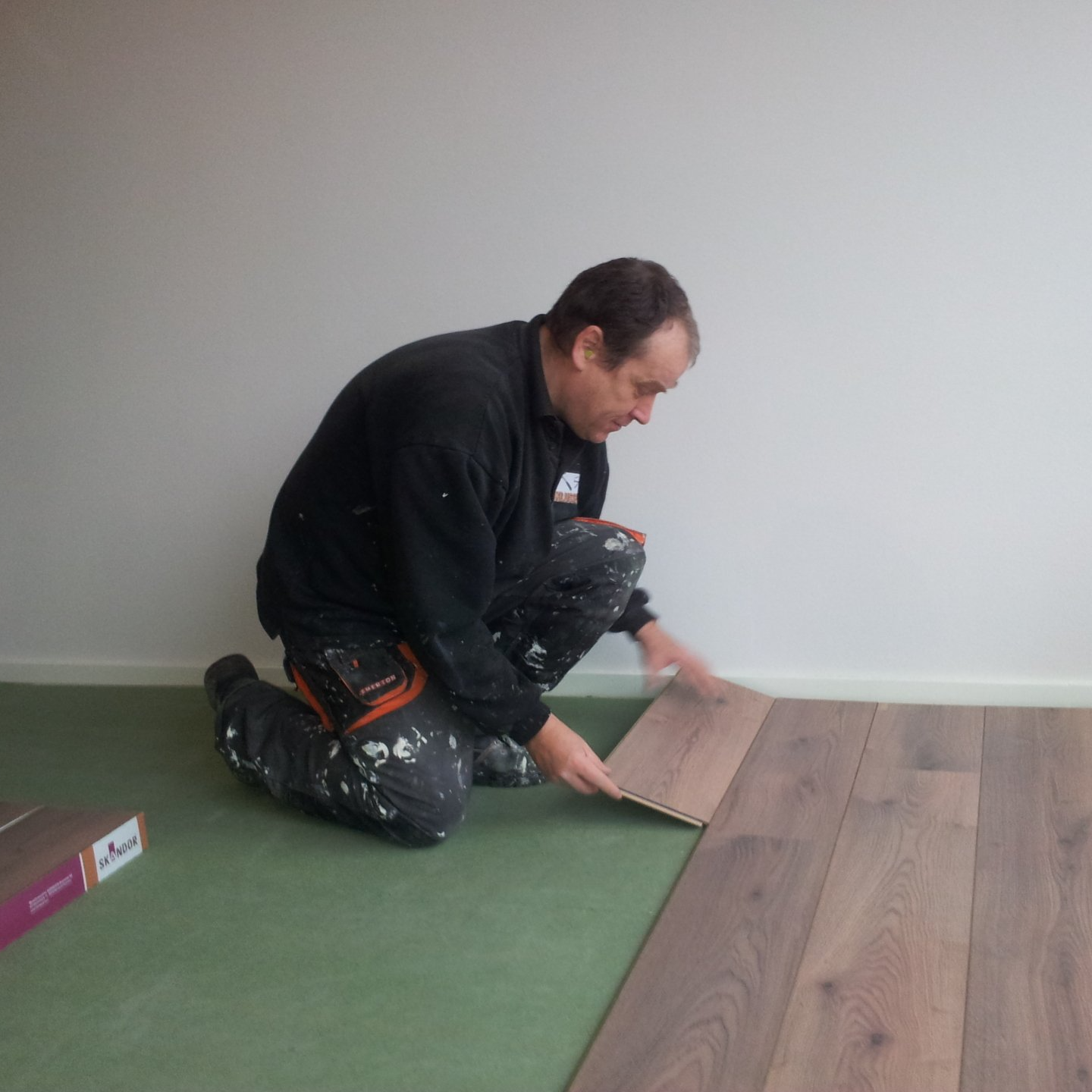 Een persoon in werkkleding knielt op een groene ondervloer en legt bruine houten vloerplanken tegen een witte muur.