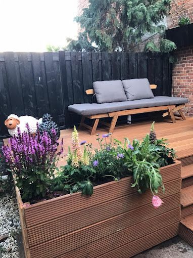Een houten plantenbak met paarse en crèmekleurige bloemen staat op een terras naast een houten bank met grijze kussens en een schaapsbeeldje.