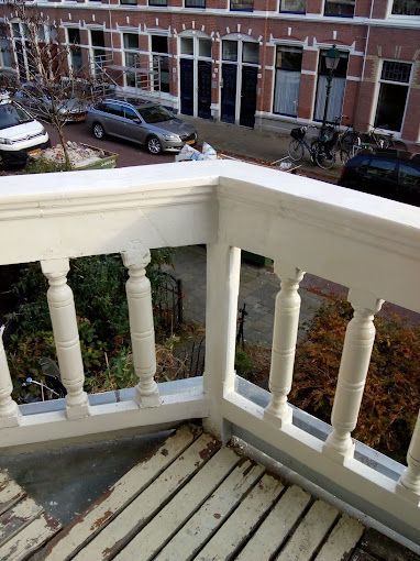 Een hoog perspectief vanaf een balkon met een witte houten balustrade, uitkijkend op een straat met auto's, fietsen en huizen.