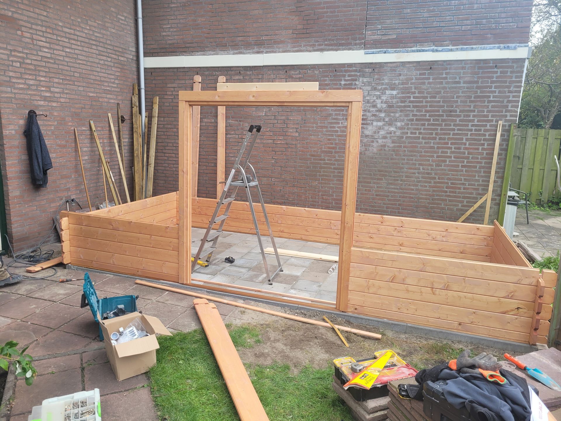 Houten wanden van een klein bouwwerkje tegen een bakstenen muur, met een deurkozijn en een trapje in het midden.