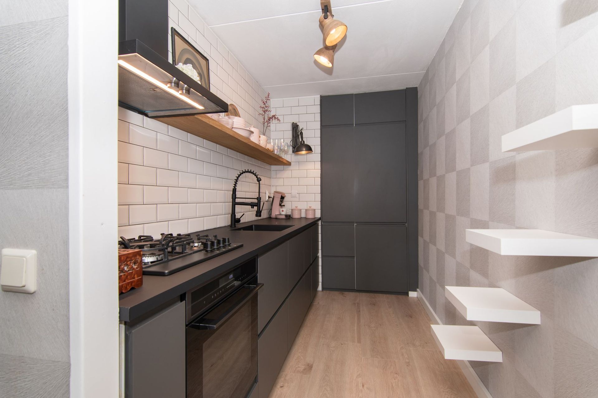 Een moderne, smalle keuken met grijze kasten, zwarte werkbladen, witte metro-tegels, een houten plank en zwevende planken.
