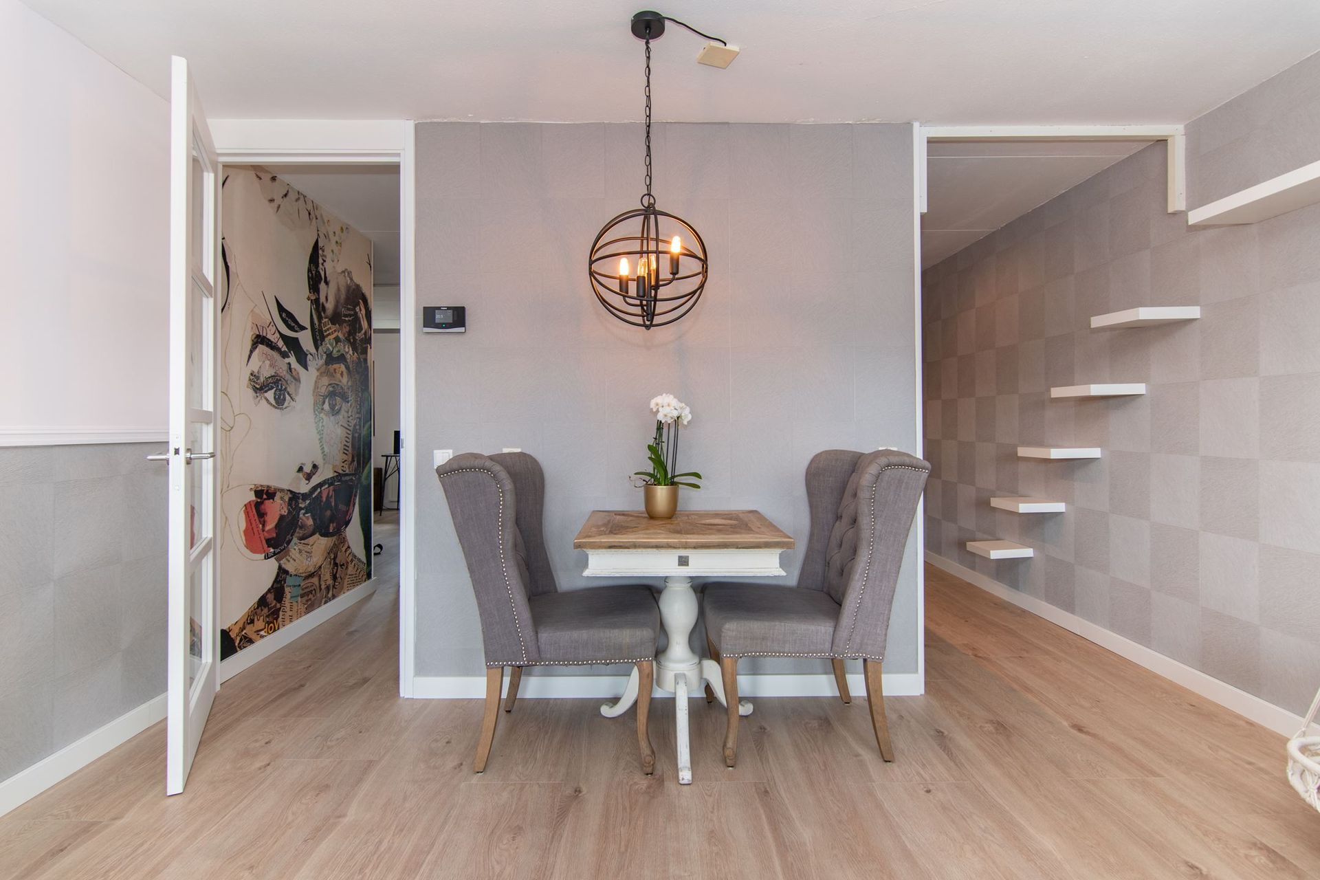 Een eethoek met twee grijze oorfauteuils en een kleine houten tafel onder een metalen bolvormige hanglamp in een moderne kamer.