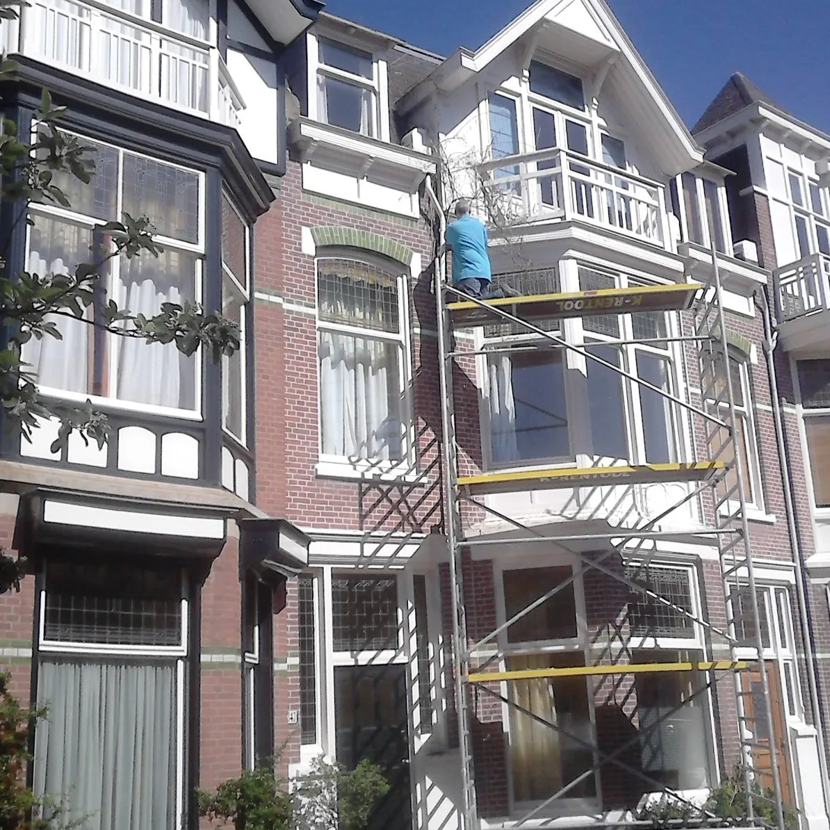 Een persoon in een blauw shirt werkt op een steiger die aan de voorkant van een drieverdiepingen tellend bakstenen herenhuis is bevestigd.