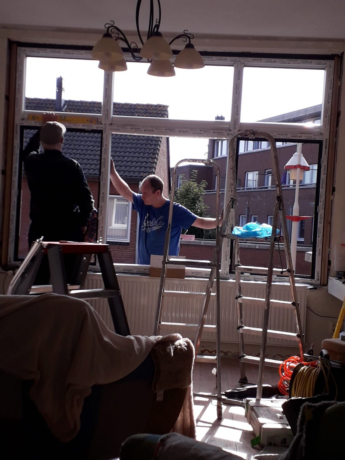 Twee mensen zijn staand op ladders bezig met het installeren of repareren van een groot raamkozijn in een kamer.