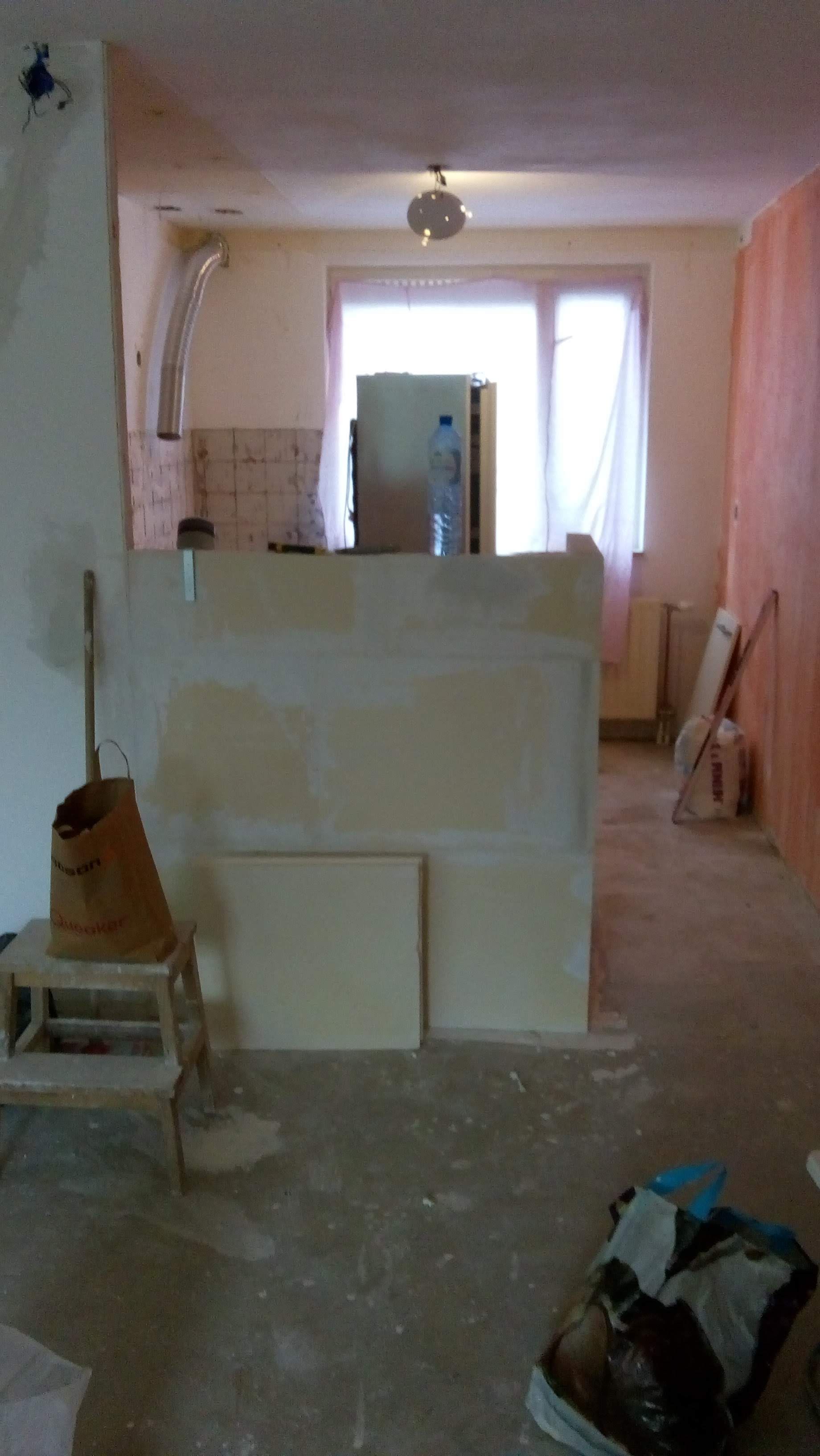 Een kamer in aanbouw met een gedeeltelijke gipsplaten scheidingswand in het midden, een krukje met een bruine papieren tas en een raam aan de achterkant.