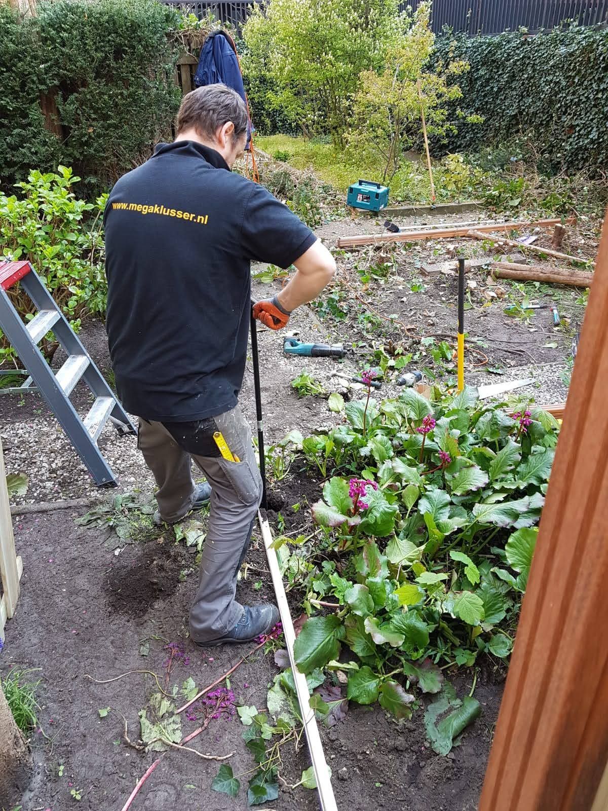 Een persoon in een zwart shirt en een grijze broek staat in een tuin en bewerkt met een werktuig de grond in de buurt van wat groene planten.