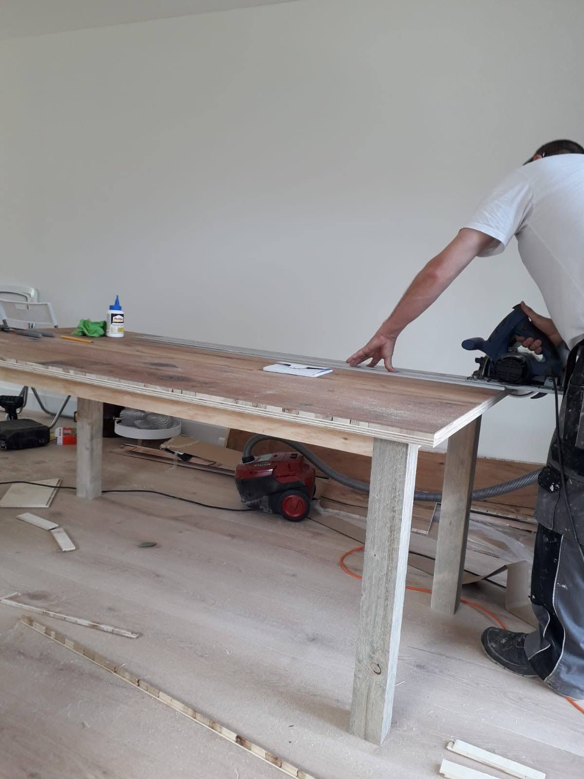 Iemand gebruikt een cirkelzaag om de rand van een grote houten tafel in een onafgewerkte kamer bij te zagen.