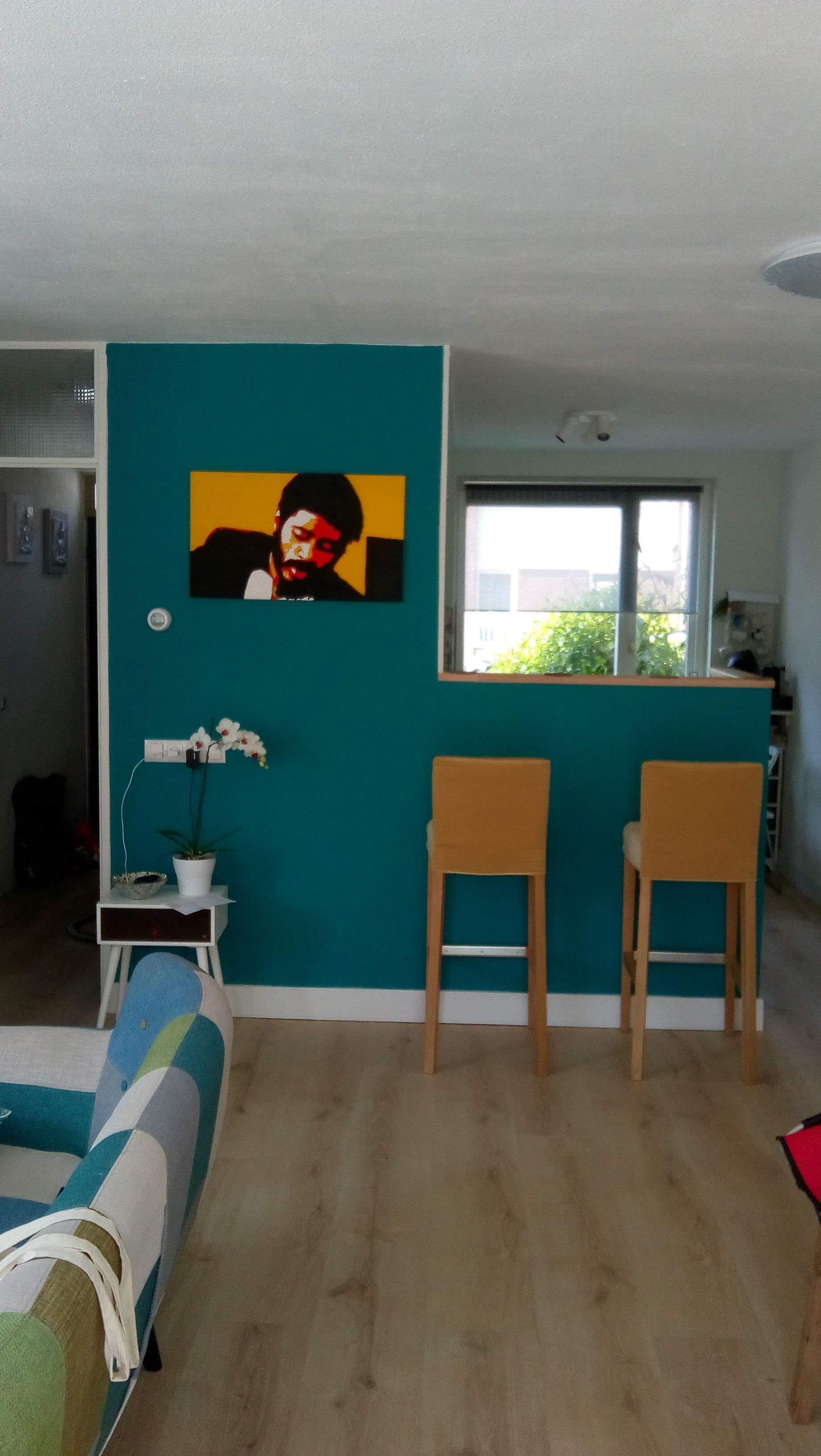 Een turquoise accentmuur met een pop-art portret, twee barkrukken, een klein bijzettafeltje met een plant en een lichte houten vloer.