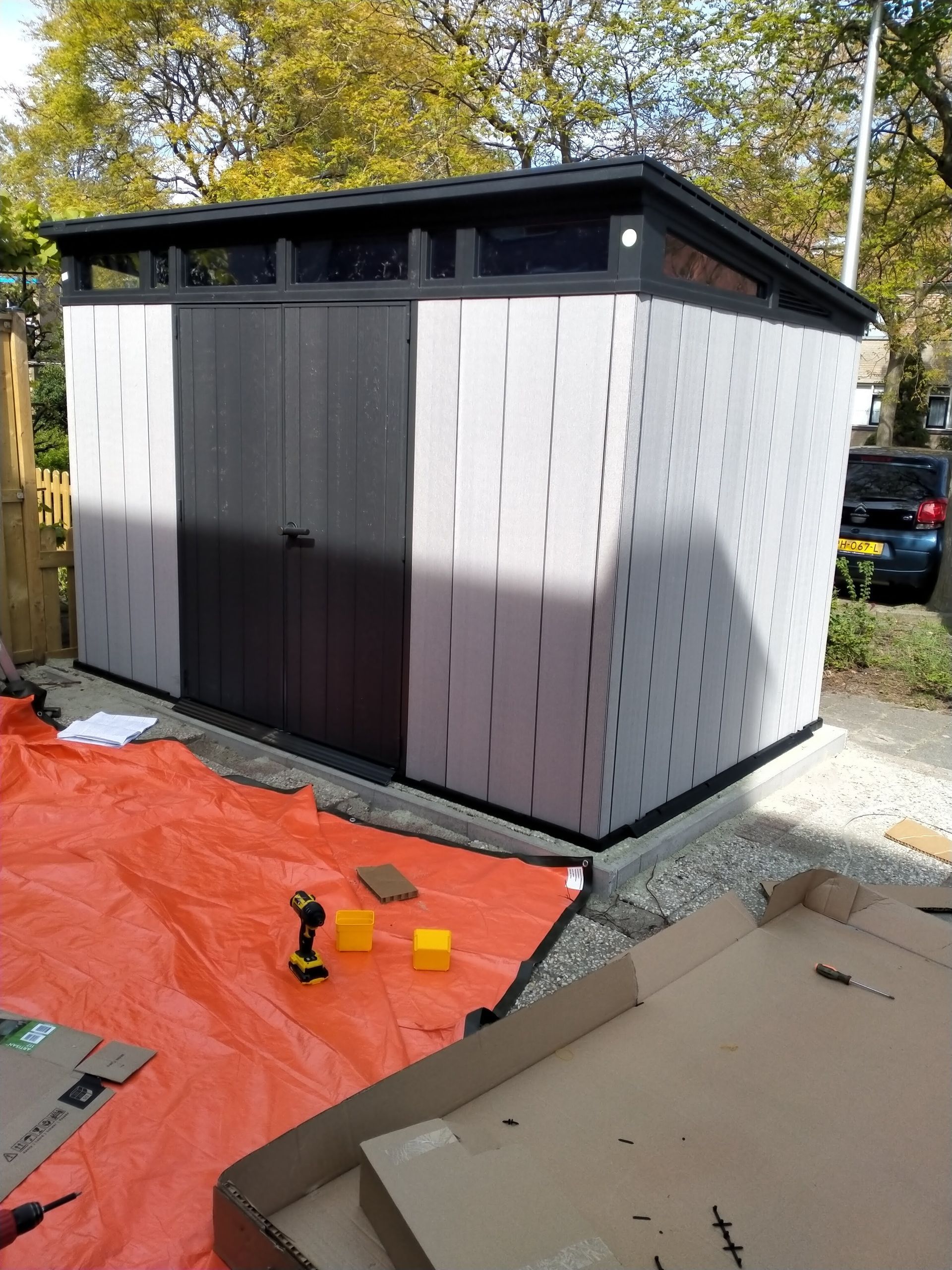Een gedeeltelijk gemonteerde zwart-grijze tuinschuur staat buiten op een grindpad, met gereedschap en materialen op een grondzeil.