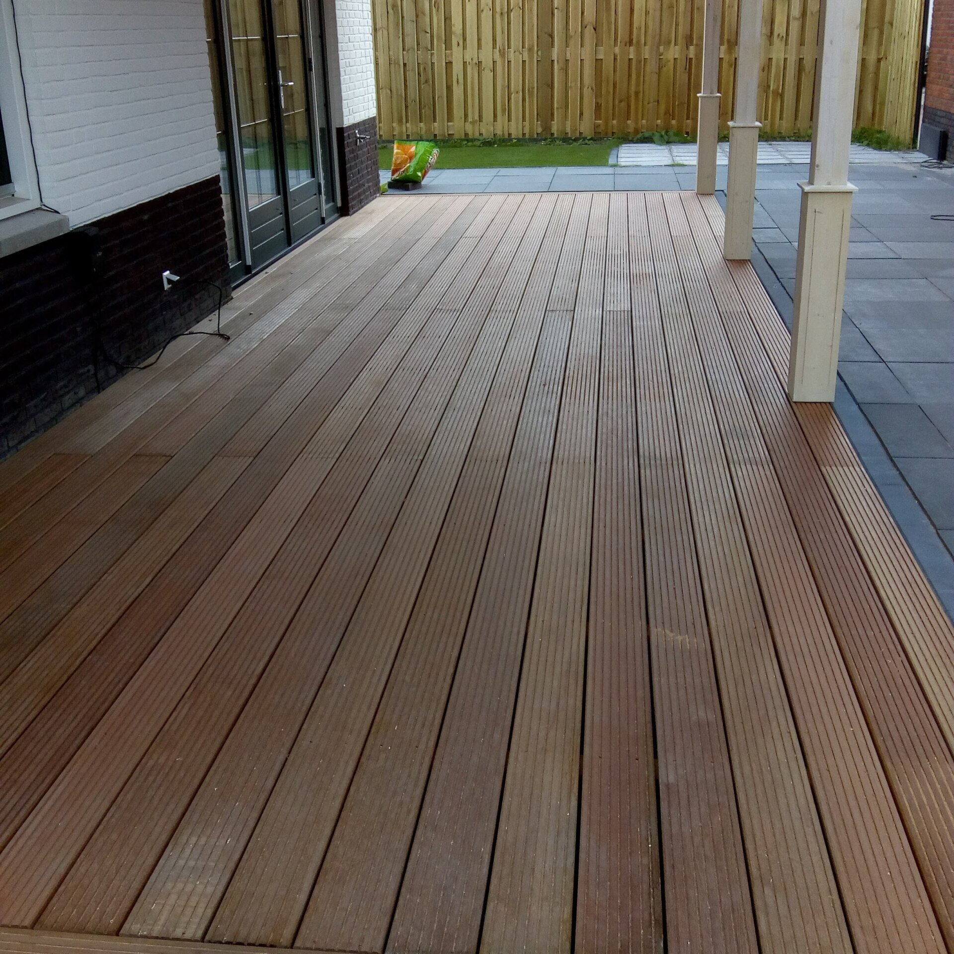 Houten terras met parallelle, geribbelde planken dat uitkomt op een patio, begrensd door een huismuur en een houten schutting.