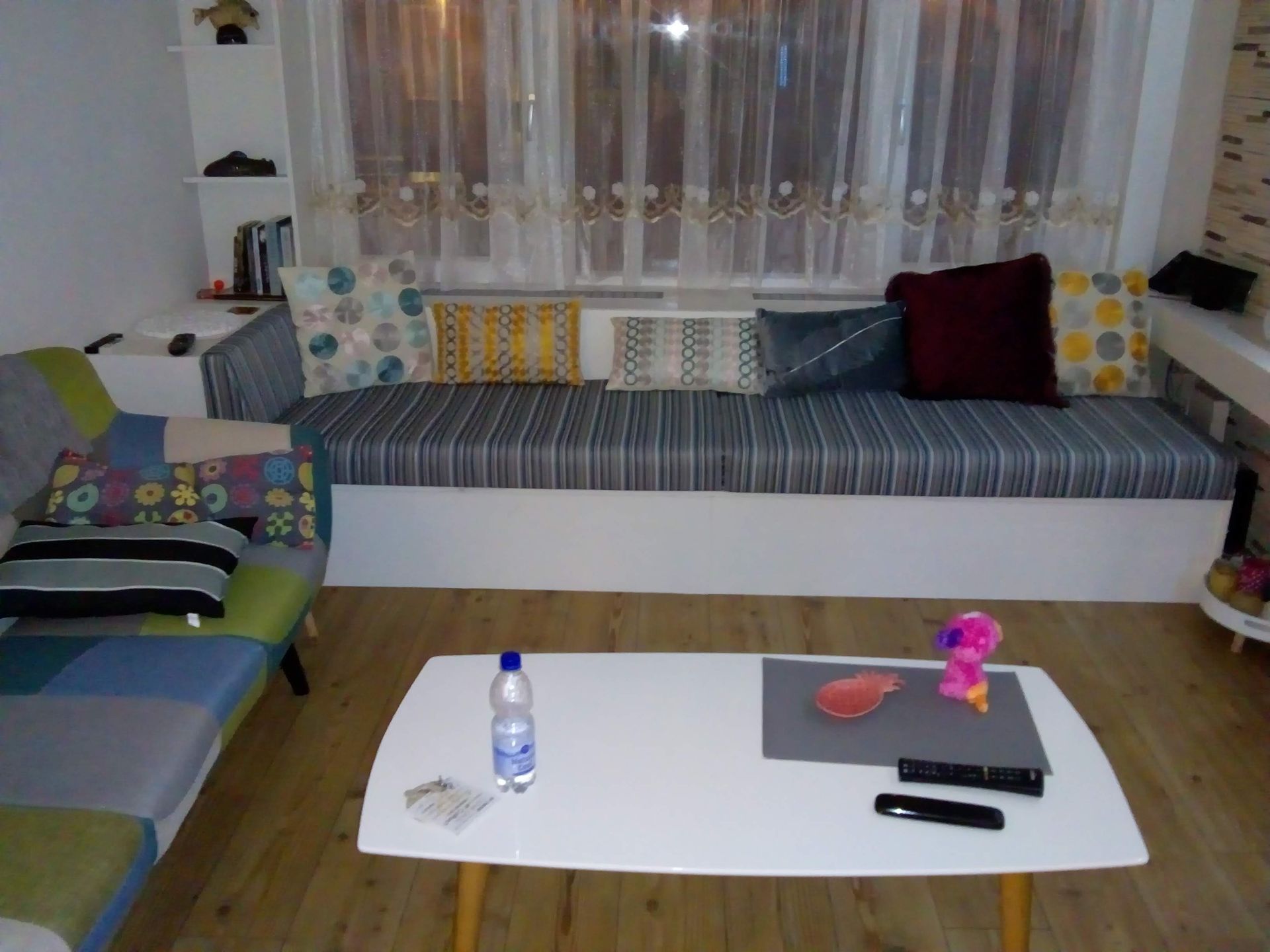 Een woonkamer met een veelkleurige bank, een witte zitbank bij het raam en een witte salontafel op een houten vloer.