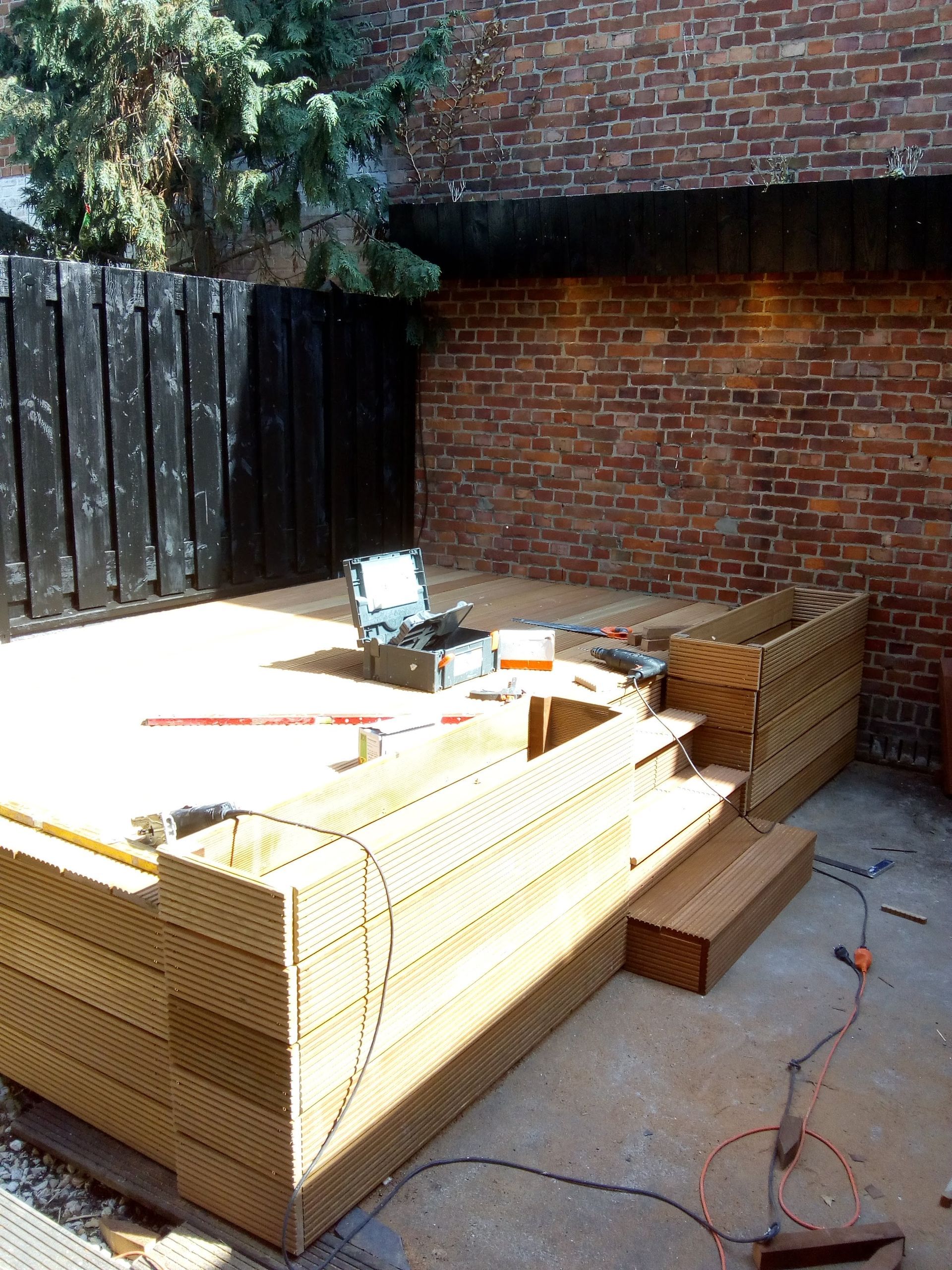 Een houten terras in aanbouw in een kleine tuin met een bakstenen muur en een donkere schutting.