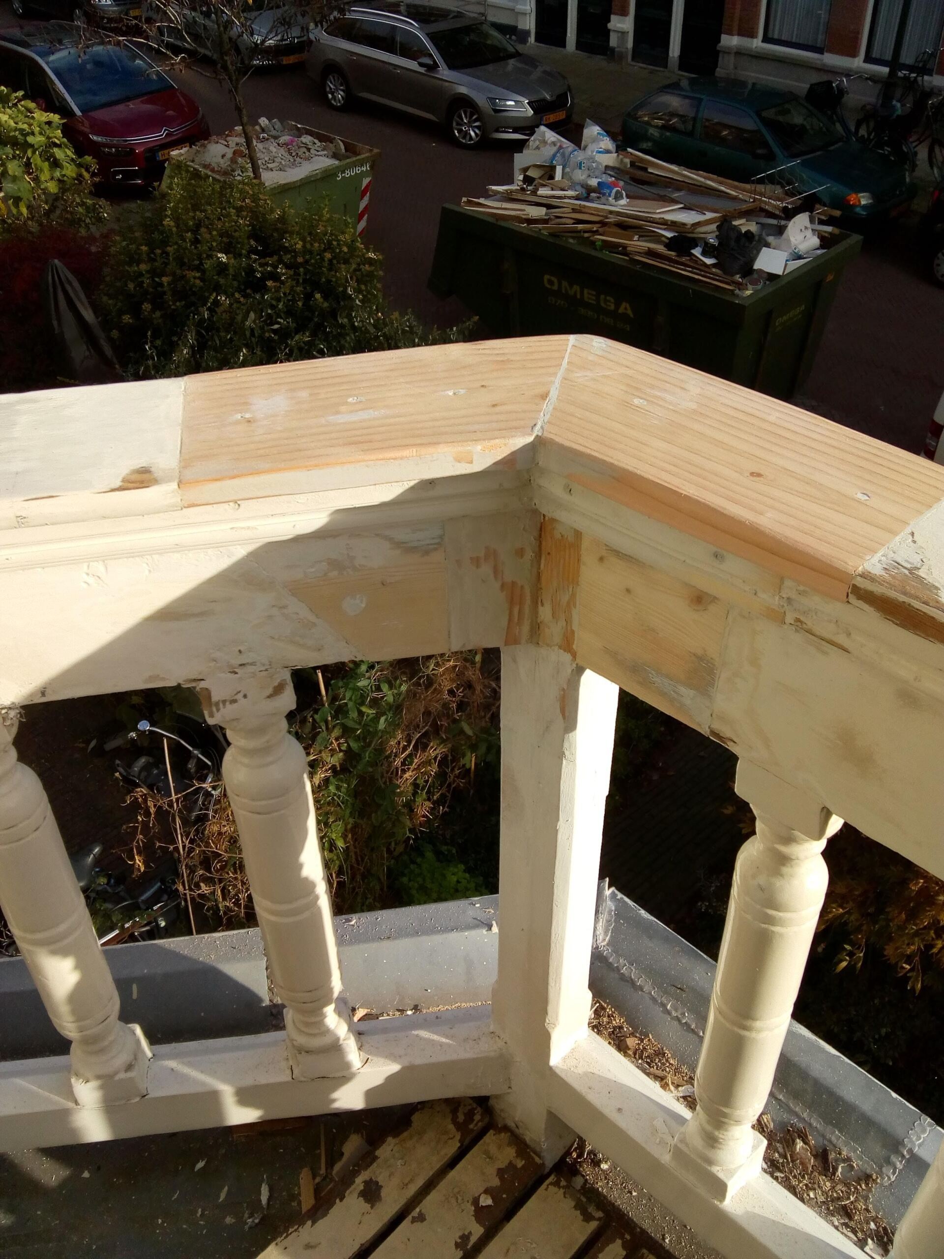 Een close-up van een balkonreling met onbewerkt houten afdekkingen en witgeschilderde balusters, met uitzicht op een straat met geparkeerde auto's.