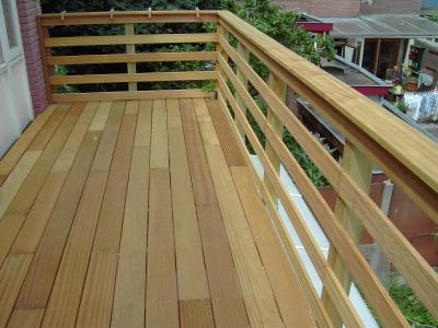 Een houten balkon met een horizontale balustrade, met uitzicht op een tuin en daken.