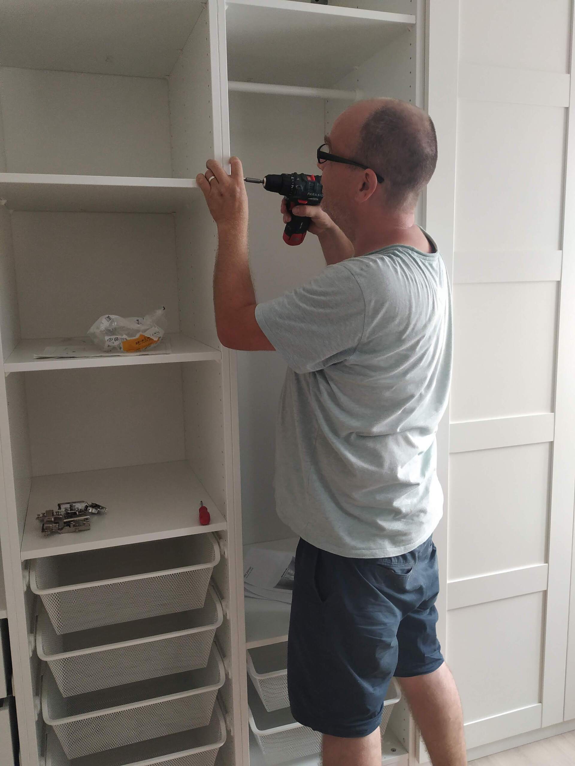 Een persoon gebruikt een accuboormachine om bevestigingsmateriaal te installeren in een wit modulair kledingkastsysteem.