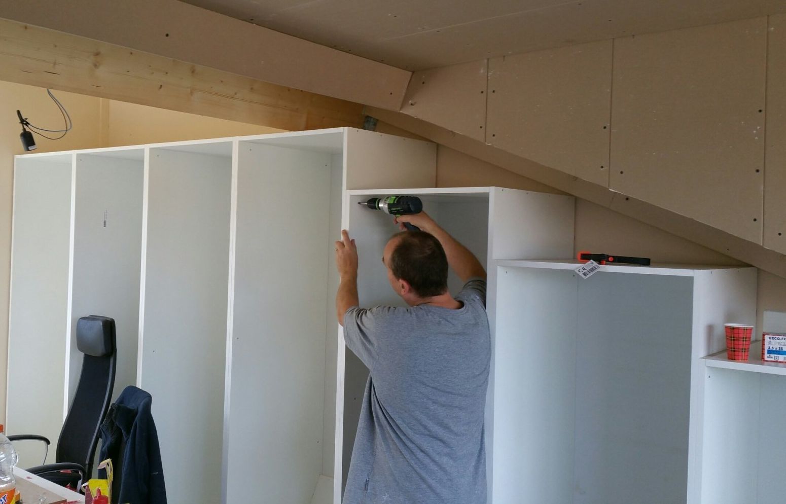 Iemand gebruikt een boormachine om op maat gemaakte witte planken te monteren tegen een schuin plafond in een ruimte die in aanbouw is.