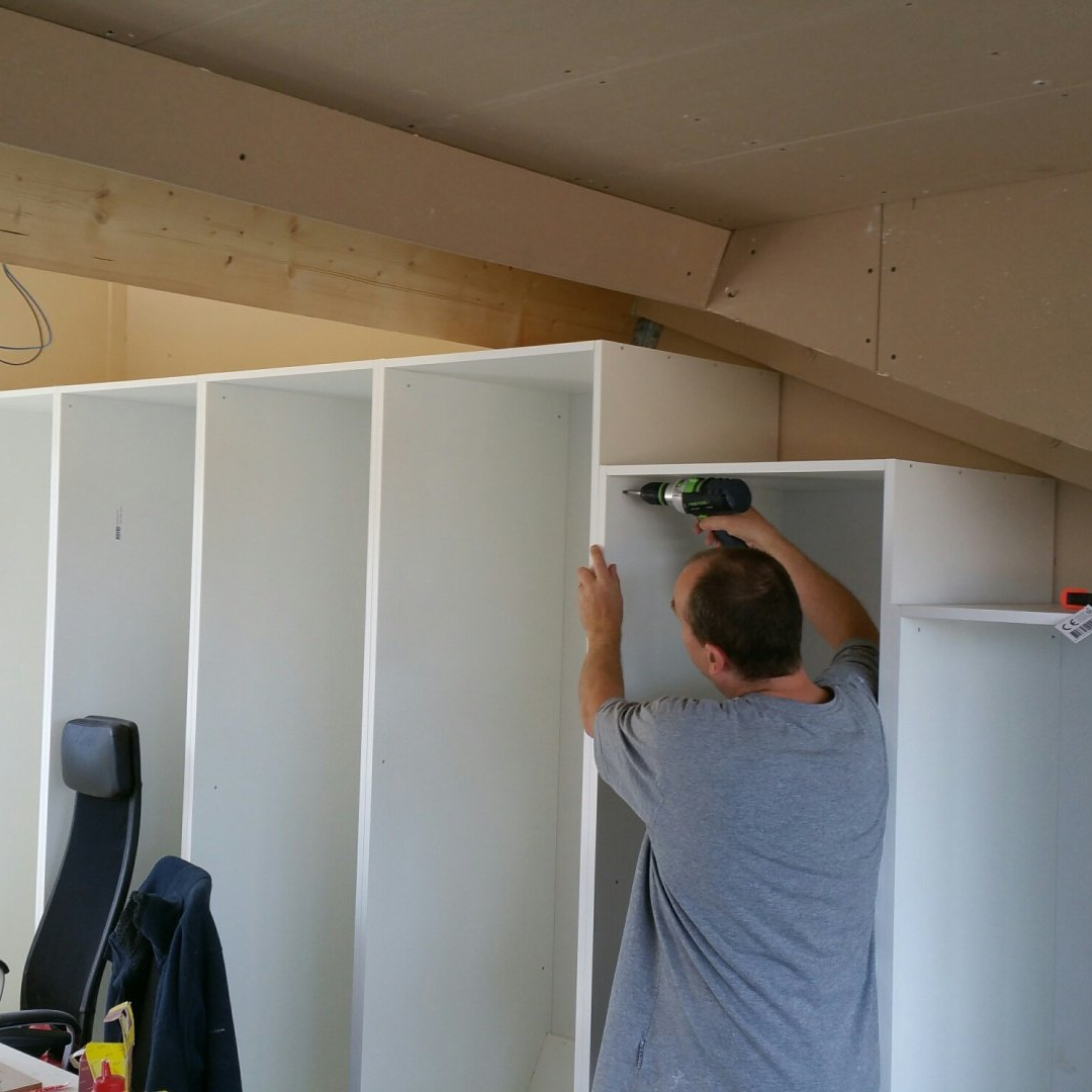Een persoon in een grijs T-shirt gebruikt een boormachine om witte houten planken in elkaar te zetten in een zolderkamer met schuine plafonds.