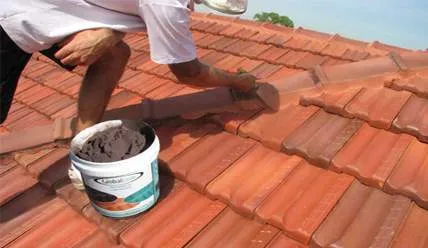 A man is kneeling on a roof next to a bucket of cement.