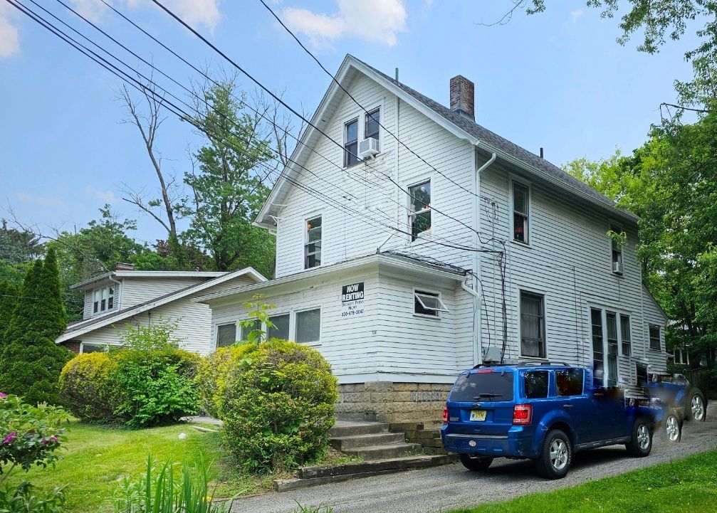 White two-story house with black shutters, stone steps, and a blue SUV parked in front