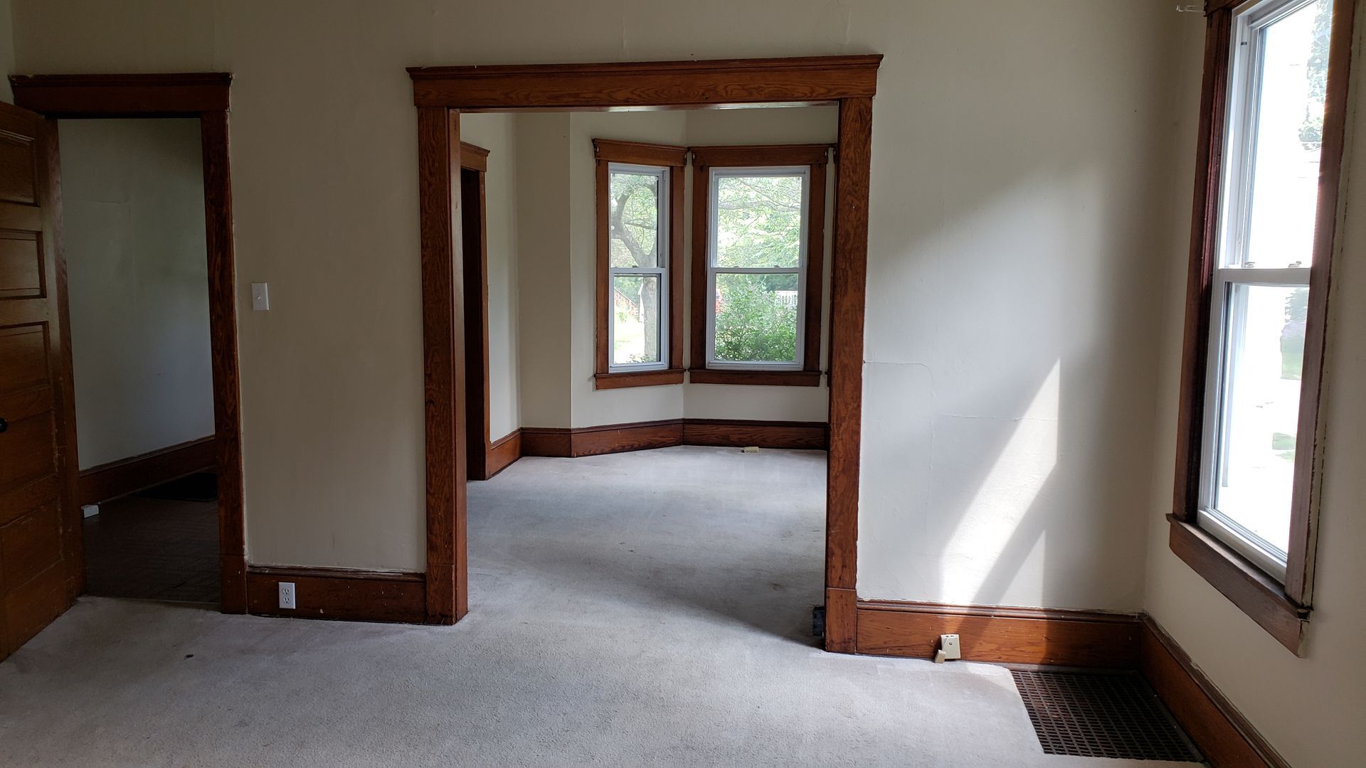 Empty room with beige carpet, white walls, dark wood trim, and windows on two sides.