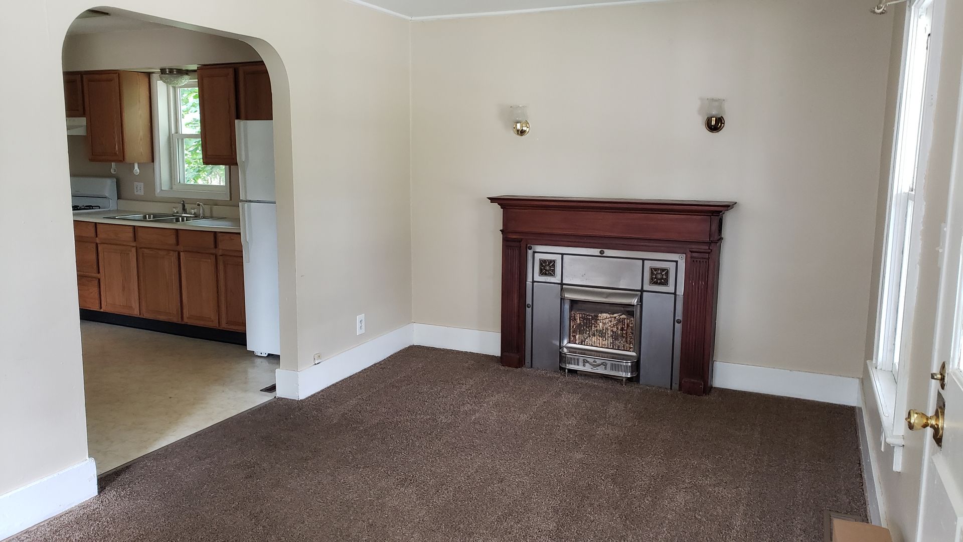 Empty living room with brown carpet, beige walls, and a dark wood fireplace mantel.