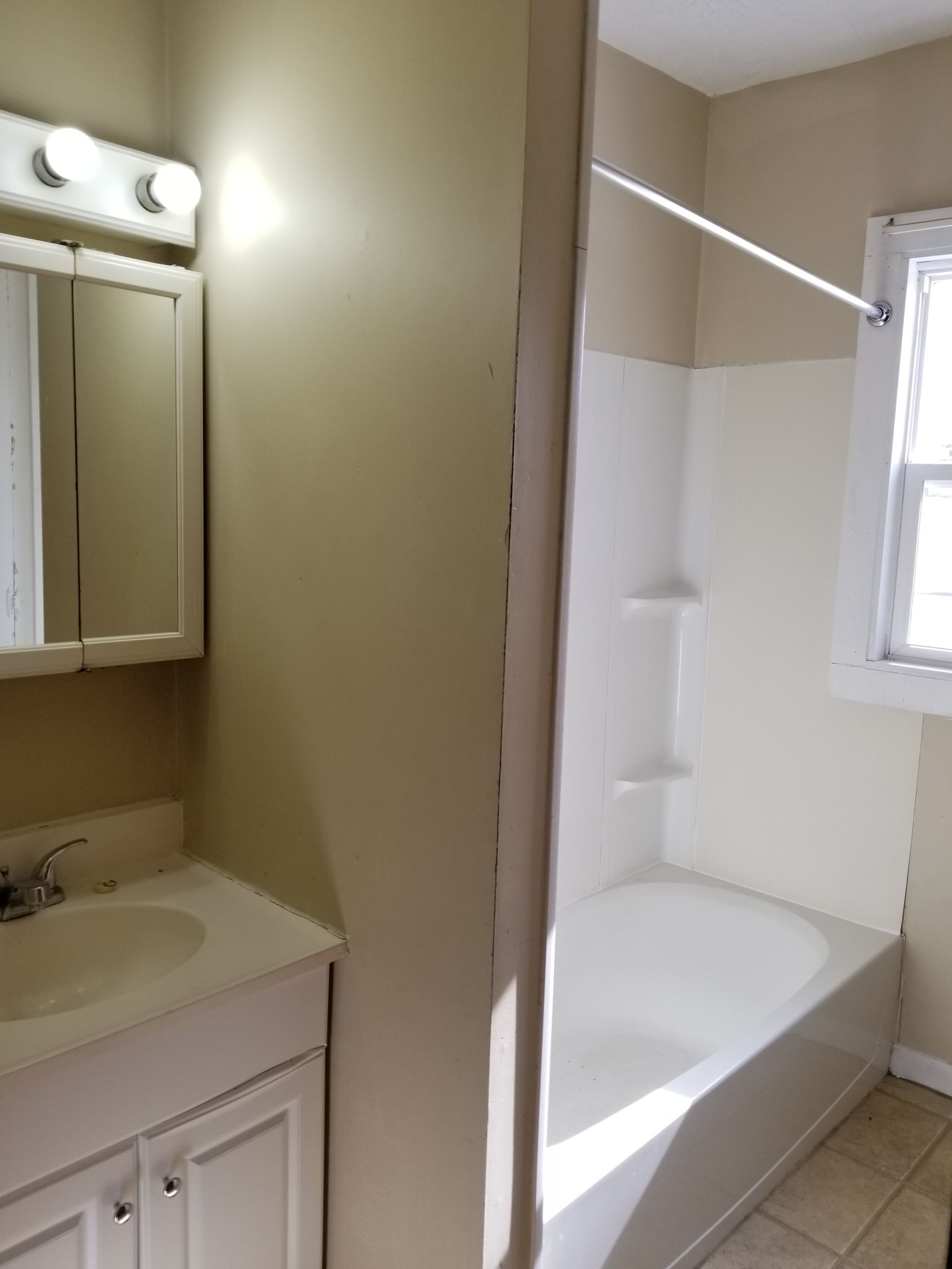 Bathroom with sink and mirror on left, bathtub and window on right, beige walls and tile floor