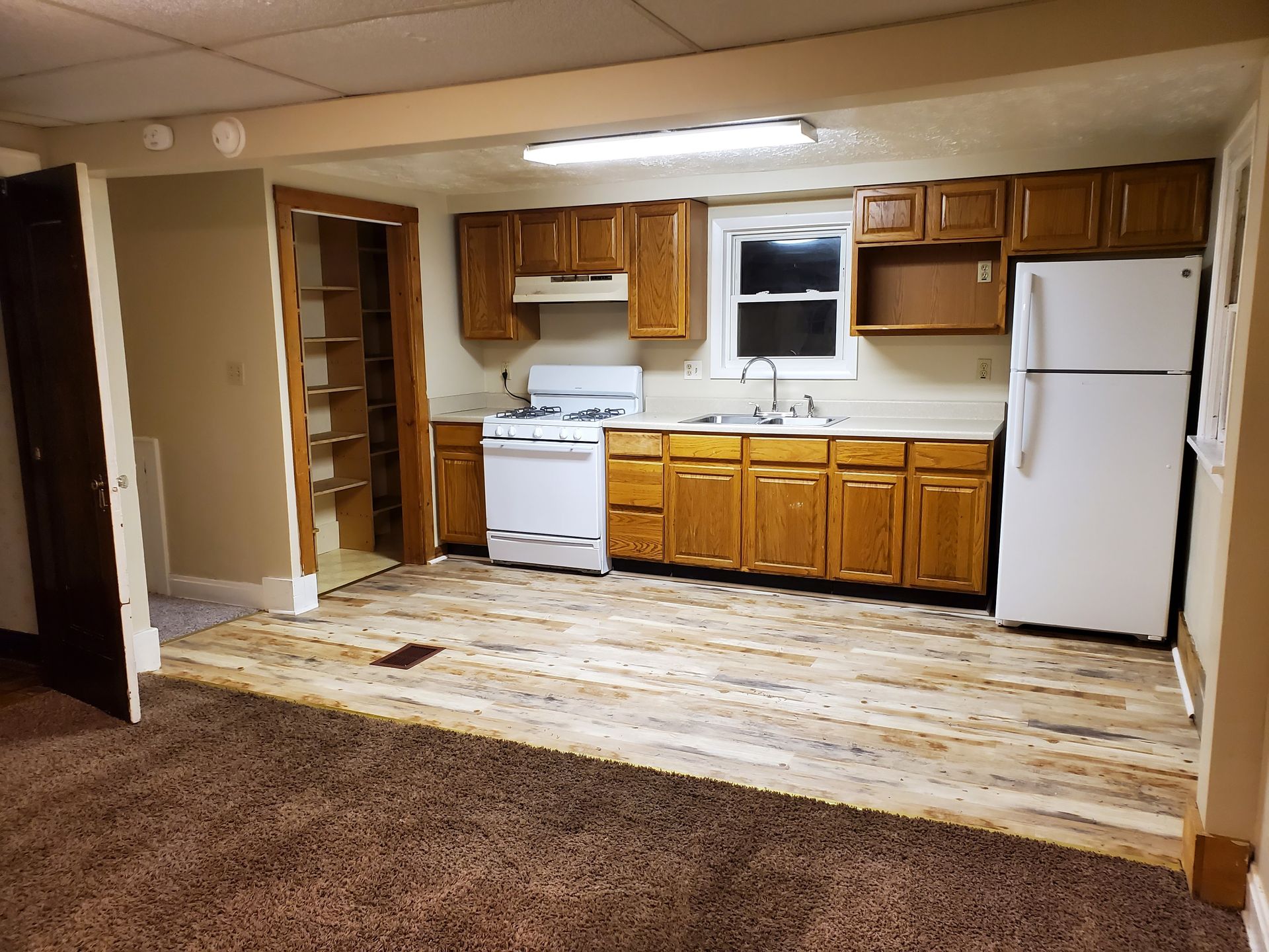 Empty kitchen with white appliances, wood cabinets, and a raised linoleum floor beside worn carpet.