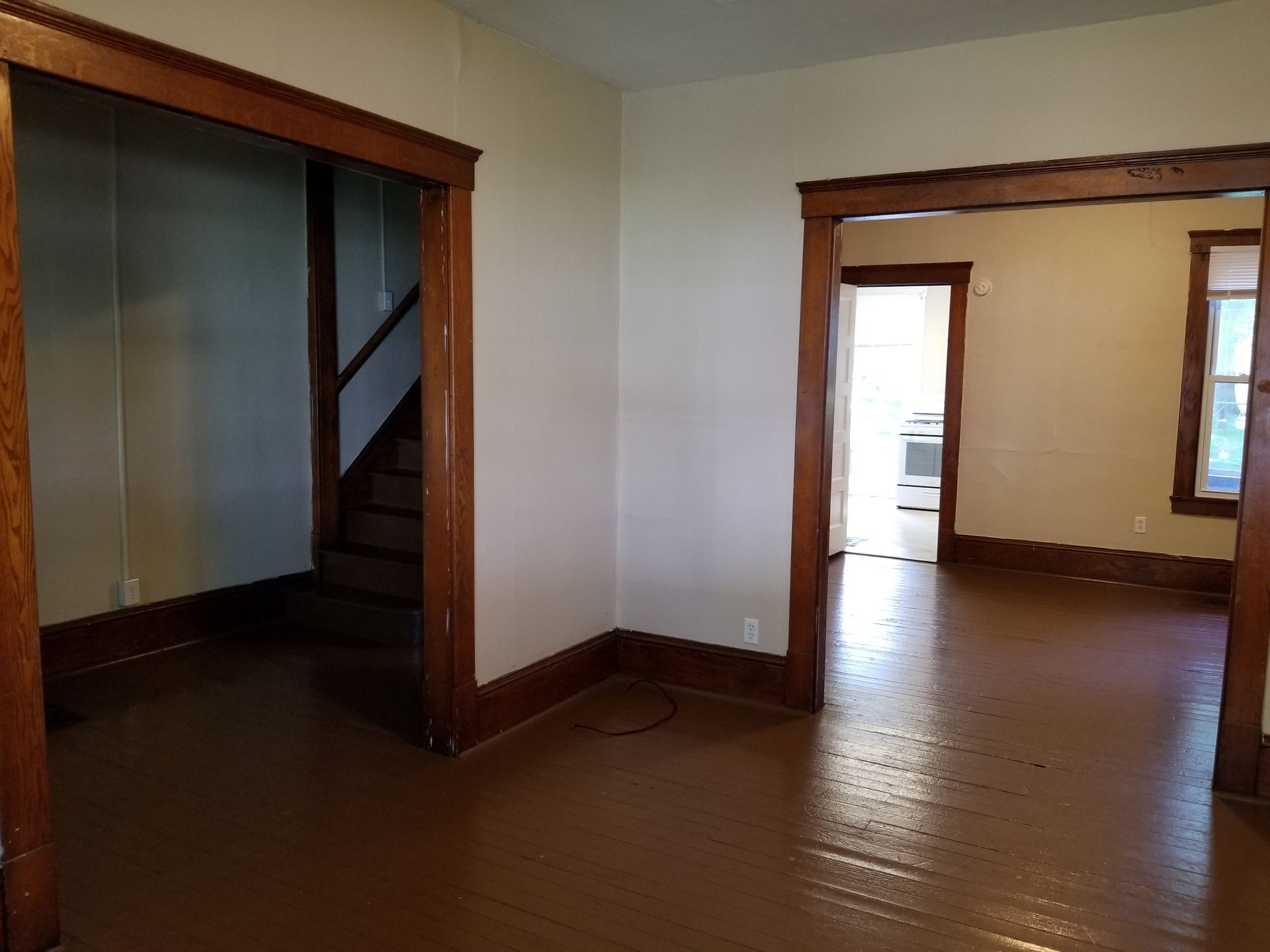 Empty interior room with hardwood floors, wood trim, and a doorway to a bright adjacent room.