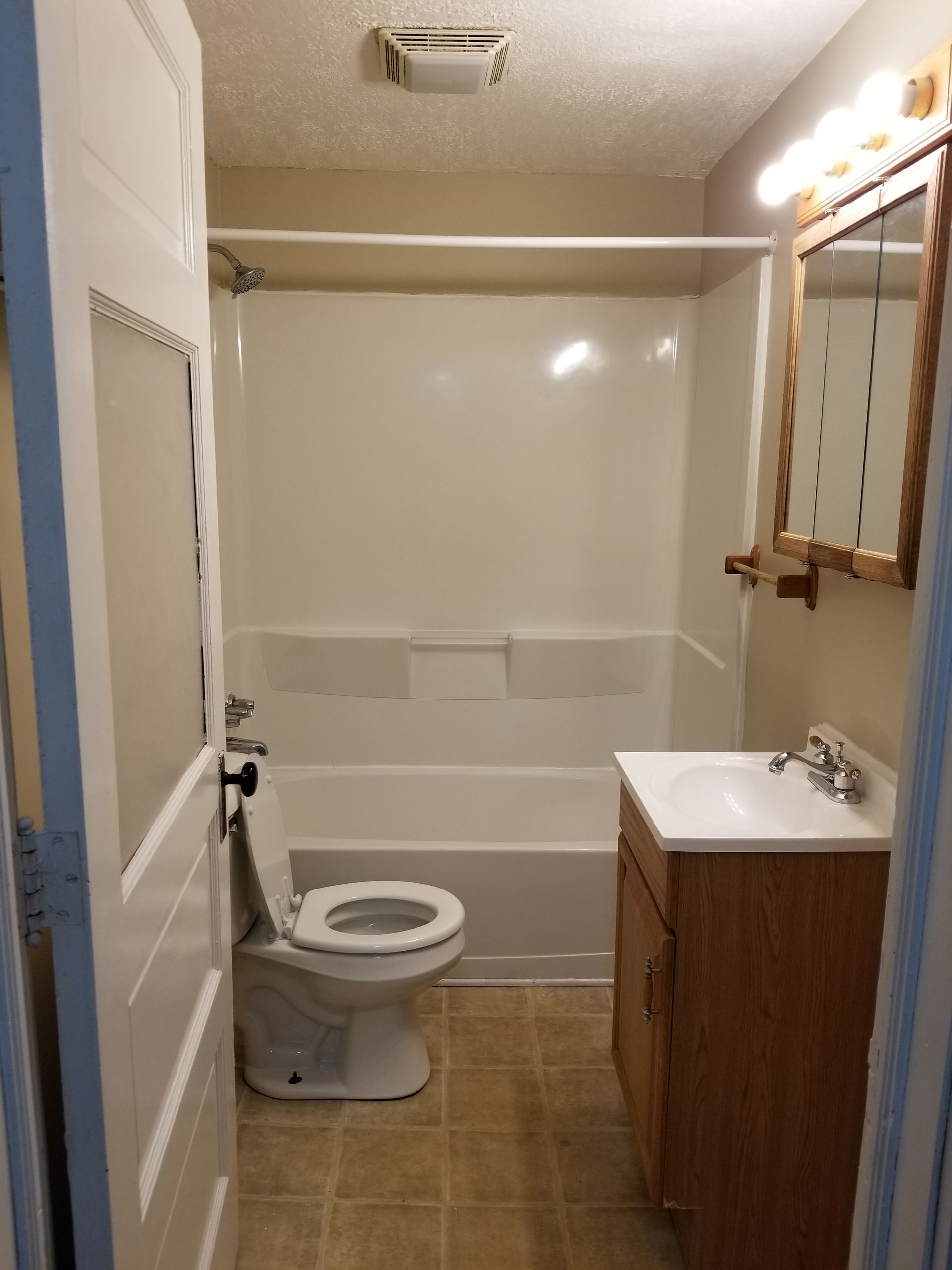 Small beige bathroom with toilet, sink vanity, mirror, bathtub, and bright vanity lights.