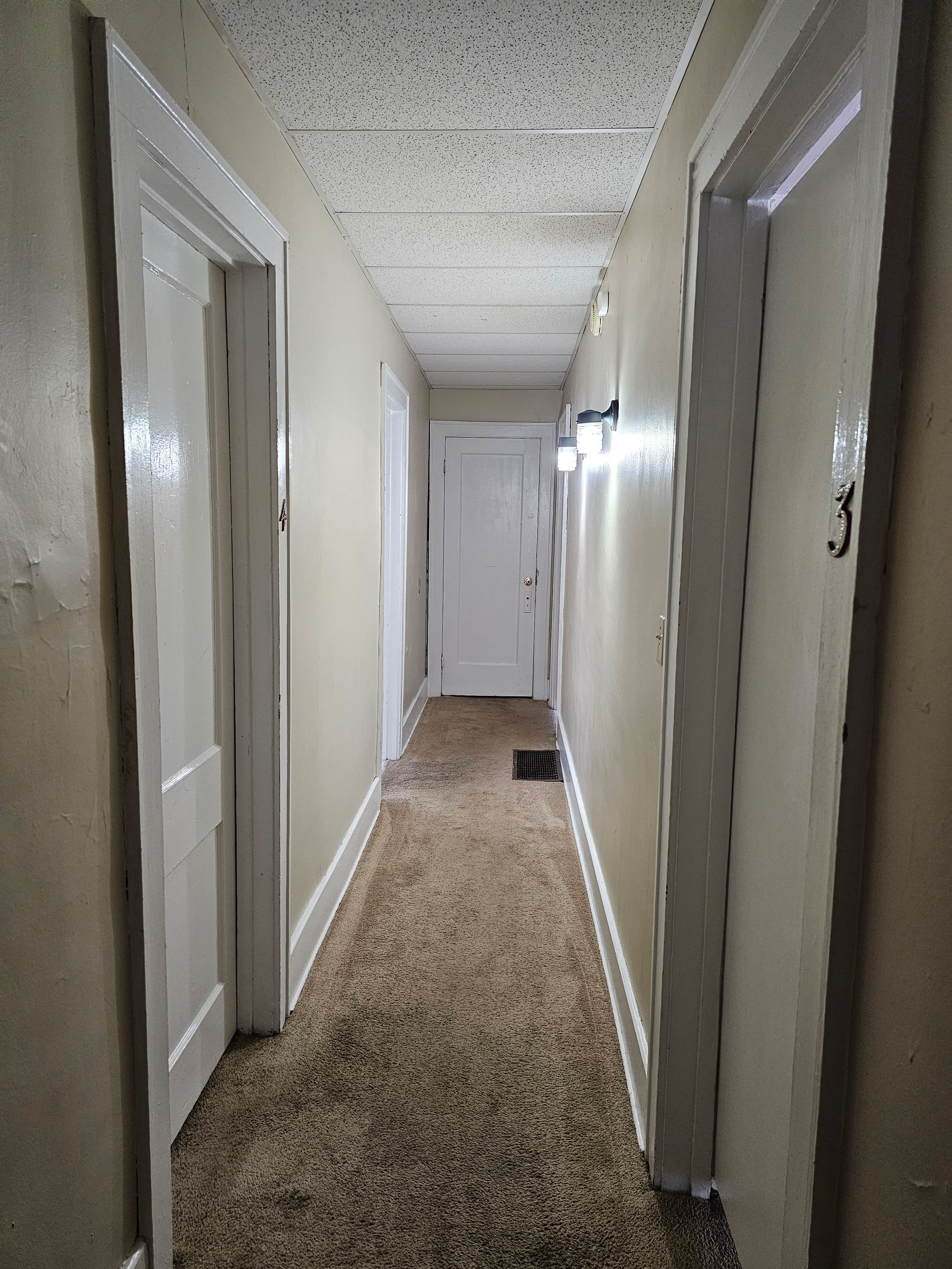 Narrow beige hallway with white doors and speckled ceiling, viewed toward a closed door at the end.