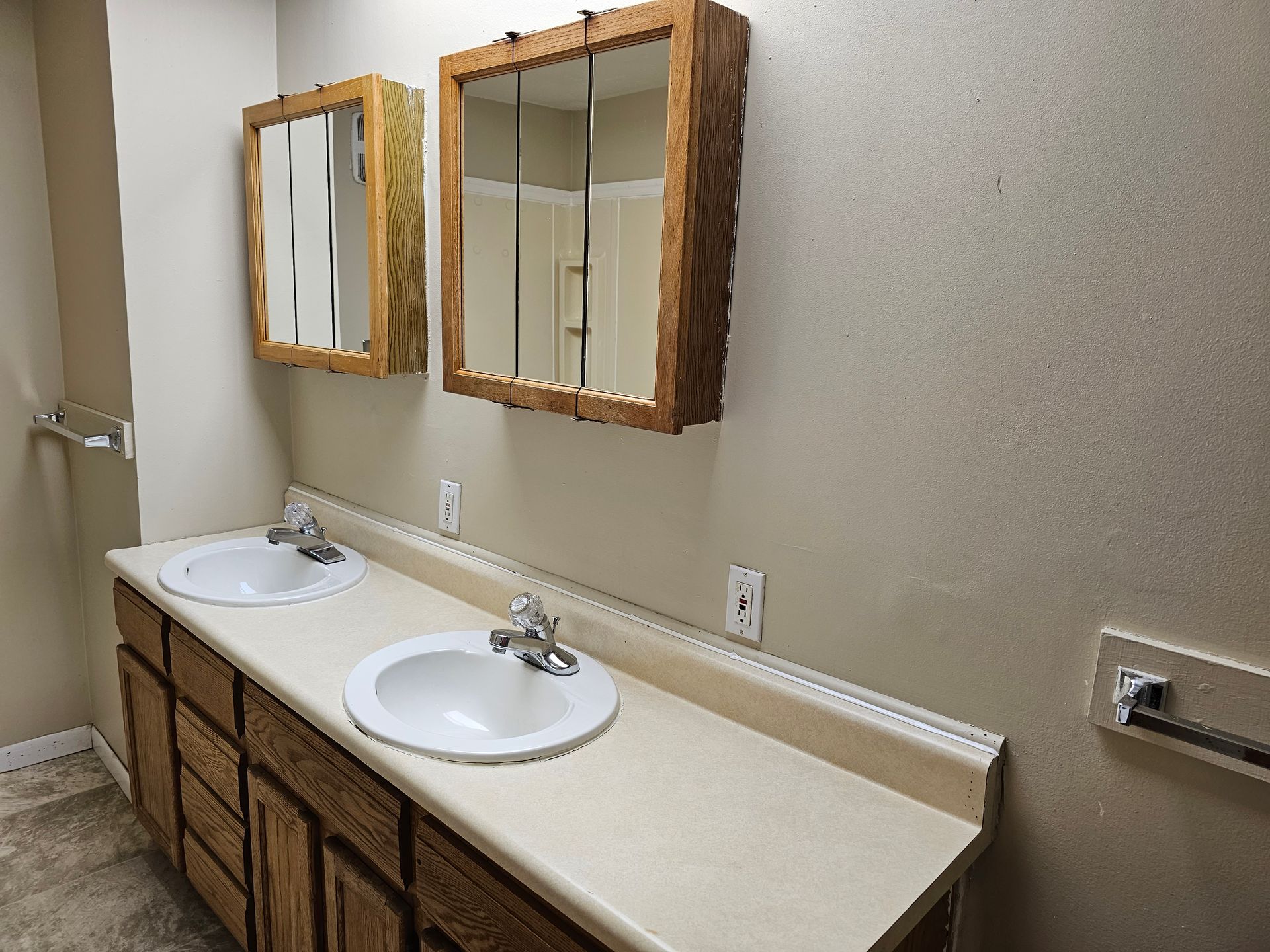 Bathroom vanity with double sinks, wood cabinets, and two mirrors on a beige wall.