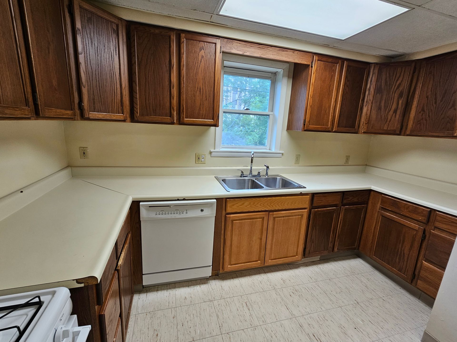 Small kitchen with wooden cabinets, white countertops, dishwasher, sink under a window, and linoleum floor.