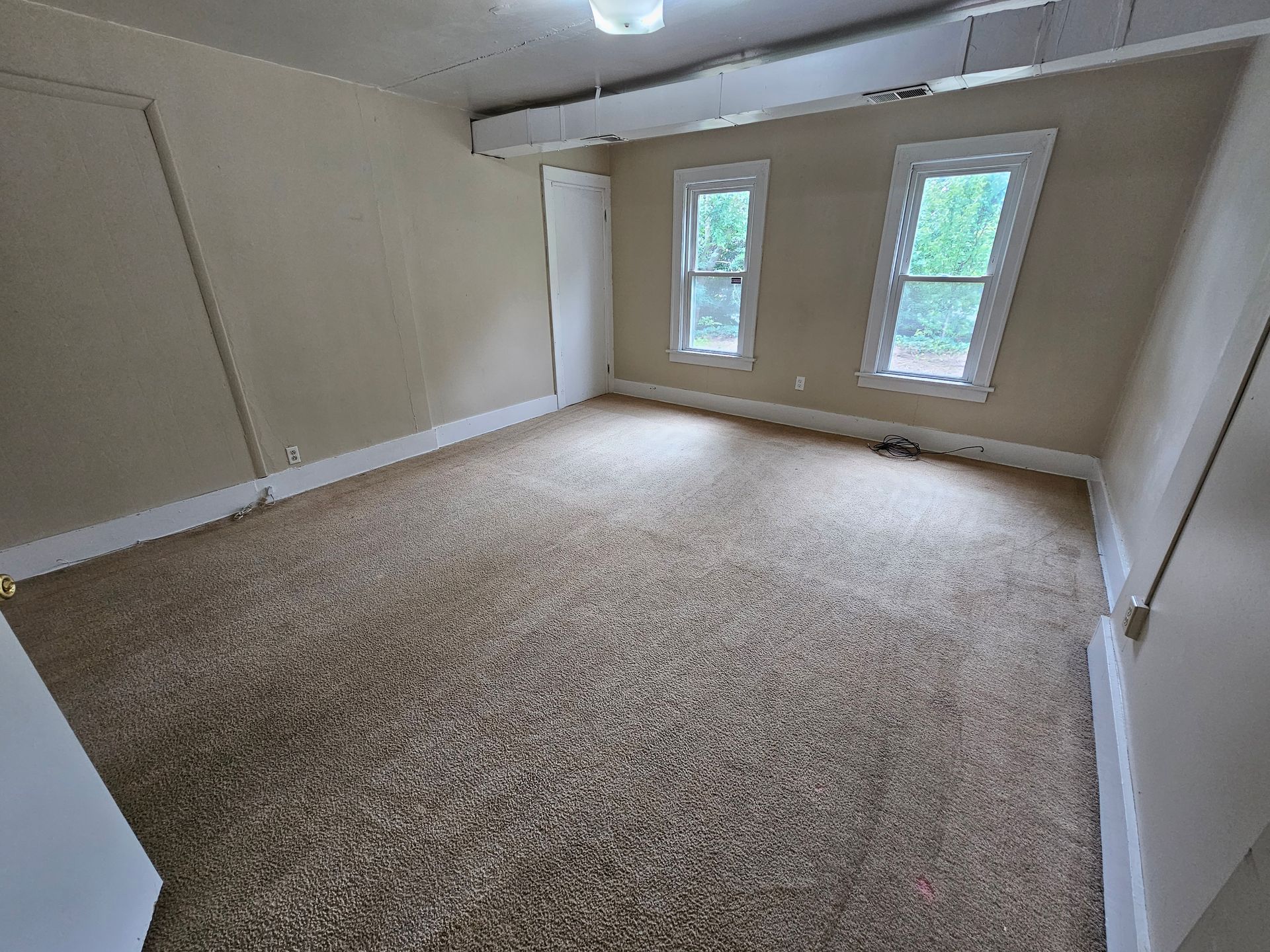 Empty beige carpeted room with two windows, white walls, and a ceiling fan.