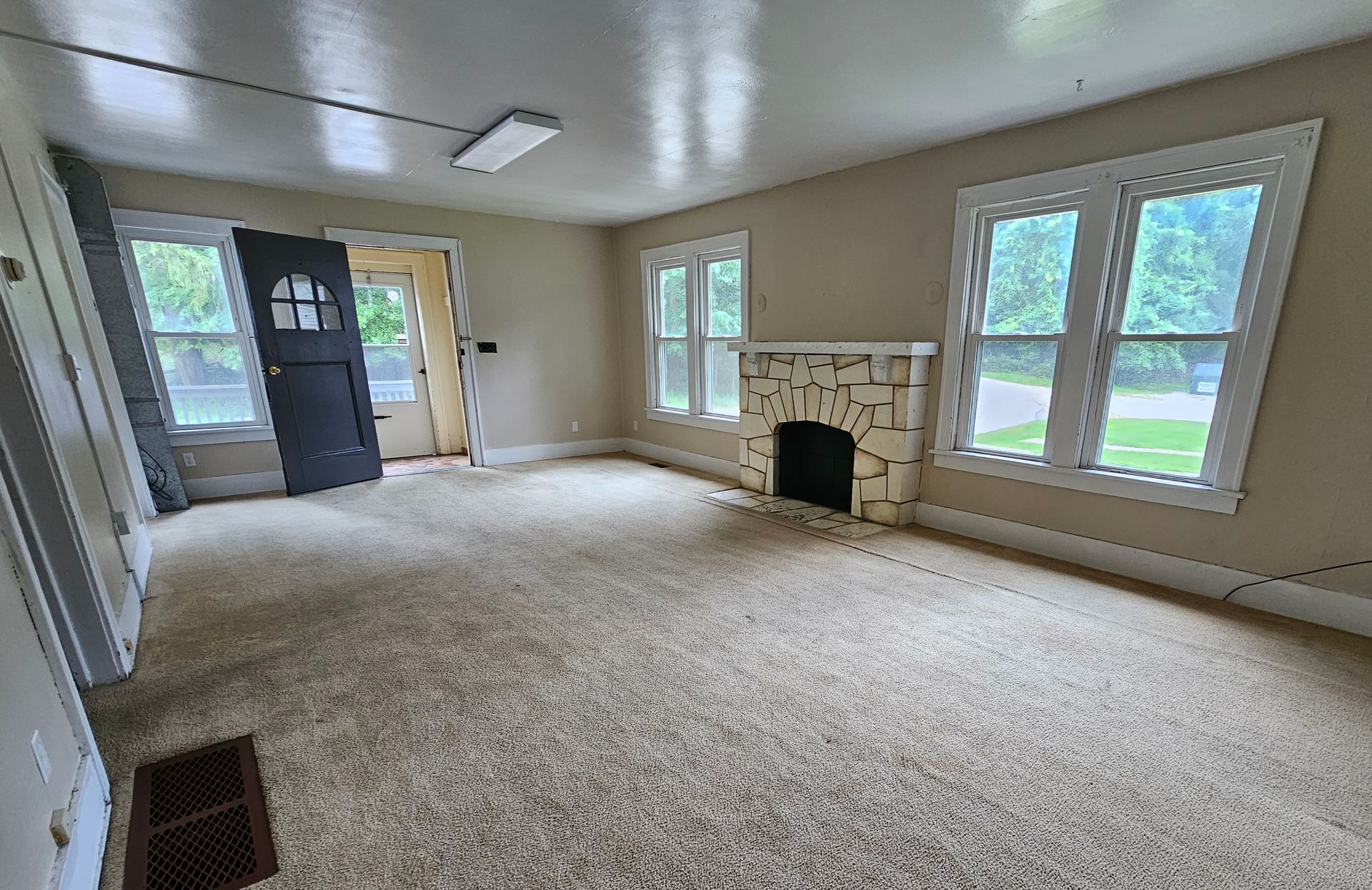 Empty beige living room with carpet, fireplace, and large windows looking onto a green yard